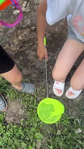 Two young children are outdoors in a grassy, wooded area. The child on the left is wearing a yellow t-shirt, black shorts, and grey Crocs. They are holding a net with a green mesh and a silver pole. The child on the right is wearing a white t-shirt with a colorful graphic, striped shorts, and a light blue baseball cap. They are holding a yellow and green bug viewer up to their eye. The child on the left appears to be handing something to the child on the right. The background shows a dirt path and a wooded hillside. The scene is lit by natural daylight.