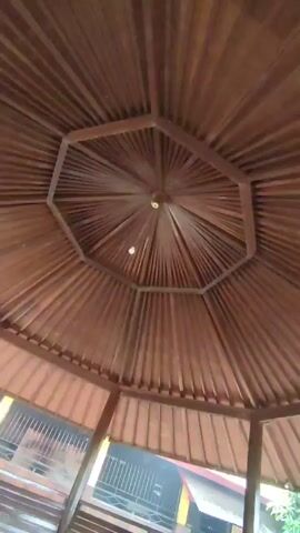 Here is a detailed description of the video:

The video shows the interior of a wooden gazebo. The camera is pointed upwards, focusing on the intricate design of the roof. The roof is made of multiple wooden slats radiating from a central point, forming an octagonal shape in the center. There is a light bulb hanging from the center of the structure. 

The wooden structure is a medium-brown color. The gazebo is open on all sides, allowing a view of the outside environment. The surroundings seem to include a building with a yellow accent and fencing. The overall lighting suggests it is daytime. The camera is relatively stable.