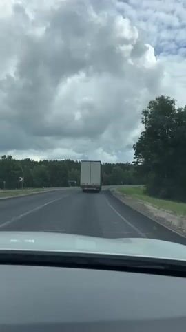 The video is shot from inside a vehicle, looking forward and slightly to the side. The vehicle is on a paved road, with a white line marking the lane. A large white truck with a tan trailer is visible ahead, traveling in the same direction. The road curves to the right. On the left side of the road, there is a patch of grass and a blue sign. On the right side of the road, there is a grassy shoulder and dense green trees. The sky is cloudy with patches of blue sky visible. The interior of the vehicle is visible in the foreground, including the dashboard and part of the side mirror. The side mirror reflects the trees and the sky. The video appears to be a recording of a journey on a highway or a country road.