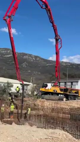 The scene is a construction site on a clear, sunny day. A large, red and yellow concrete pump truck dominates the view, with its boom extending high in the air, feeding into a construction site where workers in high-visibility vests are working within a rebar cage.  A freshly dug pit is visible in the foreground, with the rebar matrix indicating a foundation in progress. The area around the pit is sandy. In the background, there are mountains and a small, white, single-story building.  A red and white striped sign stands in the foreground, near a stack of layered materials, possibly construction lumber. The camera slowly pans to the right, revealing more of the construction site.