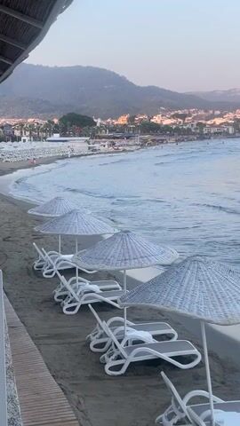 The video shows a beach scene from an elevated perspective. In the foreground, there is a wooden walkway and several rows of empty white lounge chairs with matching white woven umbrellas on the sand. The ocean stretches into the midground, with gentle waves rolling towards the shore. In the background, there's a coastline with numerous buildings and houses, backed by mountains under a clear sky. The lighting suggests either early morning or late afternoon.