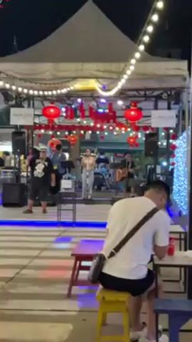 Here's a description of the video:

The video shows an outdoor venue at night, with a live band performing on a small stage. The stage is decorated with strings of lights and red Chinese lanterns. A man is sitting at a table, facing the stage, watching the performance. He is wearing a white t-shirt and black shorts, and has a satchel-type bag slung over his shoulder. There are drinks on the table in front of him.

The band consists of a female singer, a guitarist, a drummer, and a man playing some type of horn. There are a couple of other people standing or milling around the stage area. The ground is paved with what looks like zebra-striped tiles. There is a tent-like structure covering the stage area. The overall atmosphere is festive and lively. A person walks into the frame on the right side near the end.