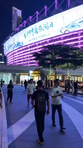 The video shows a pedestrian walkway at night with people walking in various directions. Above the walkway is a large, lit-up structure displaying the text "5.19 中国旅游日" in Chinese characters, with additional text underneath. The lighting on the structure changes from purple to yellow. A tall skyscraper can be seen in the background to the left. The area appears to be a commercial or entertainment district with shops or restaurants visible behind the people on the walkway. The general atmosphere is urban and lively.