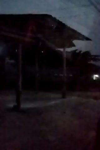 The video displays a dark outdoor scene with a rudimentary shelter constructed from wood and corrugated metal. The shelter has a sloping roof supported by posts. The ground beneath appears to be dirt or gravel. In the background, a few distant lights are visible, possibly from houses or streetlights, and silhouetted trees. The overall lighting suggests it is dusk or night. The camera appears to be stationary.