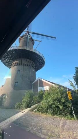 The video shows a Dutch windmill from inside a vehicle. The windmill is made of brick and has a large cap with four sails. The sails are extended and appear to be in motion. The windmill is surrounded by greenery and a few other buildings. The sky is clear and blue. The vehicle appears to be moving, as the windmill is seen from different angles. There are no people visible in the video.