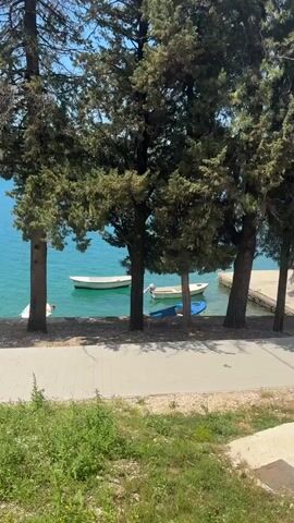 The video shows a serene waterfront scene on a sunny day. The camera pans along a paved path that runs parallel to the water's edge. The clear turquoise water is dotted with small boats, both at anchor and near a pier. Tall, evergreen trees line the path, partially obscuring the view of the water. The ground on the other side of the path is rough with exposed soil and scattered vegetation. In the distance, the path curves, suggesting a longer stretch of waterfront promenade. The sky is blue with some wispy clouds.