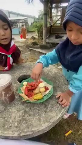 Two young girls are seated at a stone table outdoors. The girl in the foreground wears a light blue hijab and matching jacket; she reaches for food items on a green plate. The plate contains various foods on sticks, some with red sauce. The girl beside her wears a brown uniform and a brown and white scarf. Next to her sits a plastic cup with a brown liquid. The table has a hole in the center. In the background, a person in orange walks on a path, and there's a small wooden structure nearby. The environment seems to be a park or outdoor area.