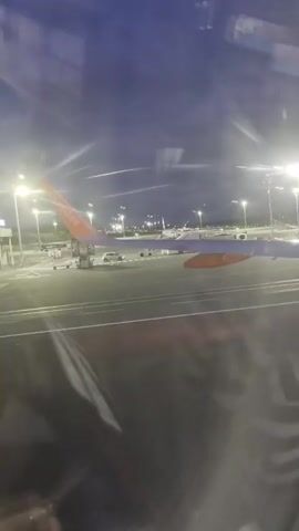 The video shows a view from inside an aircraft window. Outside, an EasyJet plane wing is visible on the right side of the frame. The airport tarmac is illuminated by bright lights. Various vehicles are present on the tarmac, including a bus, cars, and baggage carriers. The sky is dark. An announcement can be heard in the background, advising passengers to stow their seat.