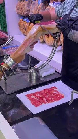 A person wearing black gloves and an apron is slicing a large piece of cured ham with a long, thin knife. The ham is secured in a metal stand. Sliced ham is arranged on a white paper on a dark countertop. In the background, many whole cured hams are hanging. The environment appears to be a food market or deli.