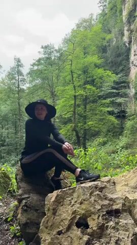 A person wearing a black hat and black athletic clothing is sitting on a rock in a forest. The person is looking to the left. The background shows a large cliff face covered in green foliage and a small waterfall cascading down the rocks. The area is lush with greenery and rocks.