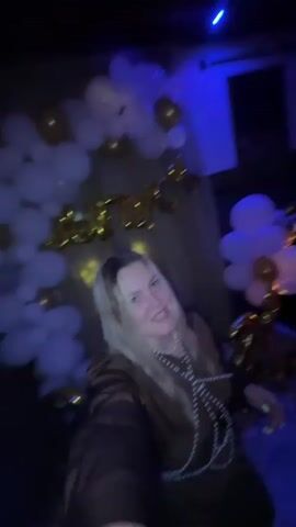 In a dimly lit room with blue and colored lights, a woman with blonde hair is captured in a selfie-style video. She is wearing a brown outfit adorned with a pearl necklace and is in the process of speaking or singing, with her mouth open in an expressive manner. Behind her, a backdrop of white and gold balloons is visible, along with large gold foil balloons that appear to spell out "ВЫПУСК," which translates to "GRADUATION" in Russian. The scene suggests a celebratory event, likely a graduation party, taking place indoors. The mood appears to be lively and festive, indicated by the woman's energetic expression and the party decorations. The time of day is not explicitly stated, but the indoor setting and lighting suggest it could be evening or night.