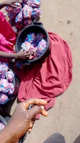 The video depicts a bustling outdoor scene, possibly a market or roadside area. The opening shot focuses on a woman in a white headscarf, crouching and presumably selling goods.  Several people are nearby, wearing various clothing.  The camera pans to show a group of people, many in white tops and purple skirts, walking on a dirt road. A motorcycle is parked nearby. The next shot shows a man working on something on a dirt lot next to a building.  The video then shifts to a paved road with a silver sedan parked at the side.  People walk along the sidewalk. Finally, the video shows a busy street with moving vehicles, including several yellow motorbikes, and more people walking. Many of the people shown are wearing red.  The environment appears to be in a developing area, with a mix of buildings, paved and unpaved roads, and general activity.