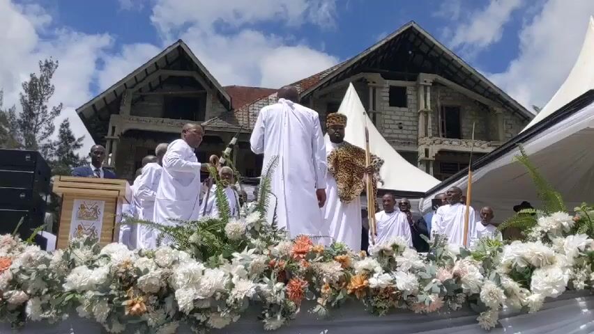 Here's a concise description of the scene:

The video shows a group of men on a stage decorated with flowers, likely outdoors due to the visible building and tent structure in the background. Most of the men are wearing white robes. One man is prominently featured wearing a leopard print garment over his white robe and holding a spear. One man is standing at a wooden podium. They appear to be participating in a ceremony or event, possibly of cultural or ceremonial significance. The sky is partly cloudy.