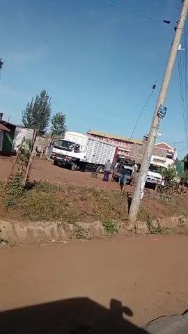 The video, shot from inside a moving vehicle, shows a roadside scene in what appears to be a rural or semi-urban area. A dirt road runs alongside a concrete ditch or curb. Several vehicles, including a white truck and a darker SUV or truck with an open back, are parked alongside the road. Buildings, some appearing to be shops or small businesses, are visible in the background, including one with a pink facade. A few people are seen walking in the area. Utility poles and wires run overhead. The sky is clear and blue. The overall environment appears dusty and somewhat undeveloped.