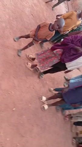 A group of children of varying ages are lined up in a row on a dirt surface. They are facing away from the camera, arranged from left to right, with a building visible behind them. The children are dressed in a variety of casual clothing, including shirts, pants, dresses, and sandals. Some children are wearing head coverings, and one holds a blue ball. The children stand relatively still. The scene is brightly lit by natural light.