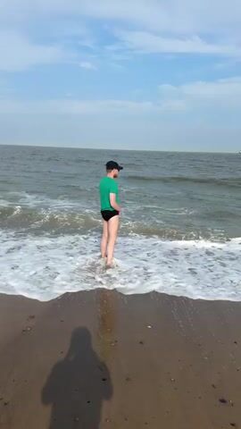 A person wearing a green t-shirt and black swimming trunks stands on a sandy beach near the water. Waves are coming onto the shore, and the person is partially in the shallow water. The person is looking down and appears to be urinating. The background shows the ocean under a cloudy sky.