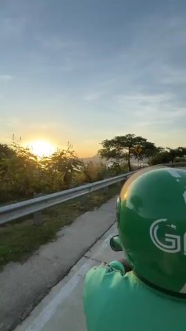 The video shows a person riding a moped along a concrete road at sunset. The person is wearing a green Grab helmet and matching green jacket. The road has a guardrail on one side, with trees and water visible beyond it. On the other side, there are trees, lamp posts, and hills in the distance. The sky is blue with some clouds. The sunset is visible in the distance over the water.