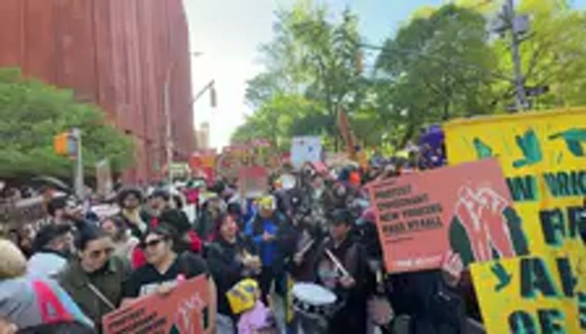 A large crowd of people is captured participating in a vibrant street protest or march on a sunny day. The scene is set on a city street, with a prominent tall, reddish-brown brick building visible on the left and lush green trees lining the right side, suggesting a park or urban green space.

Many individuals are holding protest signs and banners. In the foreground, a person on the right holds an orange sign that clearly states, "REJECT DISPLACEMENT NEW YORKERS FIGHT BACK!" with "WWW.NYCOMITE.ORG" visible at the bottom. Another large yellow banner on the far right features bird silhouettes and partially visible text that reads "NEW YORK" and "ALL OF". Other signs are scattered throughout the crowd, although their text is mostly illegible.

Participants appear engaged and energetic, with some playing drums or other percussion instruments, contributing to the rhythmic atmosphere. The crowd is moving forward, indicating a march. The audible sounds include collective chanting, which frequently repeats "Venceremos" (Spanish for "We will win"), accompanied by the rhythmic drumming. The overall impression is one of collective action, solidarity, and vocal expression against a perceived injustice or demand for change. The urban setting and nature of the demonstration align with a protest taking place in a major city like New York, United States.