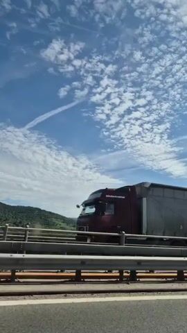 The video shows a view from a vehicle moving along a highway. The sky is blue with white, fluffy clouds, and a contrail from an aircraft is visible. In the foreground, a highway barrier with metal railings and orange accents is prominent. Beyond the barrier, rolling green hills and mountains form the backdrop. The road surface in the immediate foreground is visible, indicating the perspective of someone in a moving vehicle. No people are visible in the video. The scene appears to be a daytime shot in a rural or mountainous area.