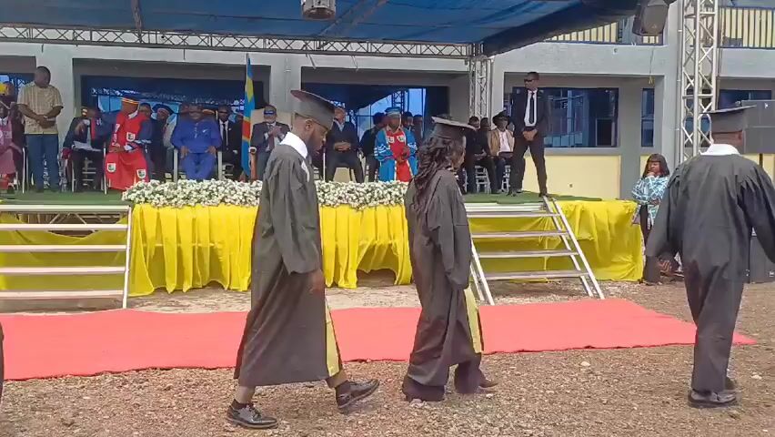 Graduates in black gowns and mortarboard hats are walking across a red carpet toward a stage. The stage is decorated with yellow fabric and white flowers, and has a backdrop with the flag of the Democratic Republic of Congo. People are seated on the stage, including individuals in suits and traditional attire. One person is speaking at a podium on the side of the stage. The event appears to be an outdoor graduation ceremony, with a blue tarp overhead.