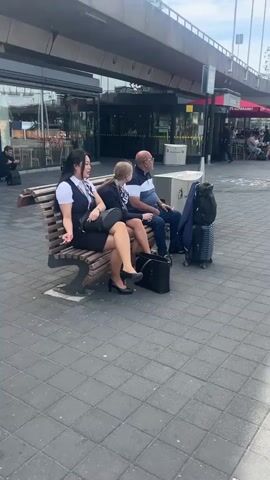 The video shows a static shot of a public outdoor space. Two women dressed in similar stewardess-style uniforms are seated on a park bench. The woman on the left is Asian, wearing a white blouse, a dark vest, a matching skirt, tan stockings, and black heeled shoes. She is smoking a cigarette, and a black purse rests on her lap. Next to her is a blonde woman in a similar outfit, consisting of a white blouse, a dark blazer, a matching skirt, tan stockings, and black heeled shoes. Her hair is pulled back in a ponytail. To the right of the women, an older man is also seated on the bench. He is wearing a white and blue striped polo shirt, blue jeans, and glasses. He is also smoking a cigarette. A black suitcase and a blue jacket are placed on the bench next to him. A trash receptacle stands behind the bench. The ground is paved with square grey tiles. In the background, there is a large building with glass windows, and some pedestrian traffic can be seen. A sign is visible on the right.