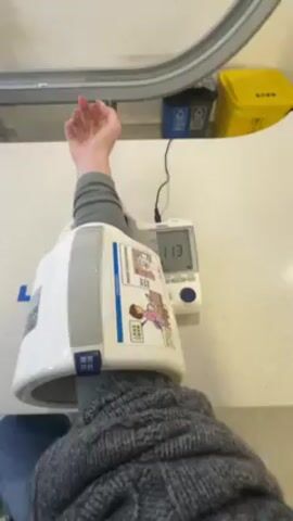 A person is sitting at a white countertop and has a blood pressure machine attached to their left arm.  The machine has a digital display that shows the blood pressure reading decreasing from 113 to 75 during the video.  The person is wearing a gray sweater.  Behind the person, there is a blue recycling bin and a yellow trash can.  There is also a curved white structure running horizontally above the countertop.