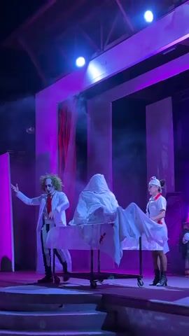 The video captures a theatrical performance with a spooky, Halloween theme, illuminated by purple and blue stage lights. In the center of the frame, on a raised platform, a person dressed as Beetlejuice, characterized by green, spiked hair, a white lab coat over a striped suit, and a red tie, stands to the left. He gestures with his left hand. Beside him, on a rolling medical gurney draped with white fabric, is a figure concealed beneath a white sheet, its shape suggesting a head and torso. To the right of the gurney, a person dressed as a nurse, wearing a white uniform with red trim and a nurse's cap, stands facing the covered figure. The backdrop consists of large, white, abstract geometric structures and red curtains. Smoke or fog machines add to the atmospheric effect, creating hazy light shafts. The overall impression is of a magic trick or a macabre scene being presented on stage.