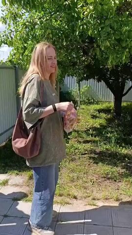 The video shows an outdoor scene with people and a house. In the first frame, a young woman with blonde hair, wearing a green t-shirt and jeans, is holding a plastic bag with food items. She is smiling and looking to her right. Behind her is a tree with green leaves and a white fence. The second frame shows a woman from behind, wearing a black tank top and yellow shorts. She has dark hair pulled back and is facing away from the camera, with her hands behind her back. She is standing in a grassy area with trees. In the third frame, a young man in a light-colored jacket and dark pants is visible in the background, standing near a green awning and some trees. The house has a light yellow exterior and a green roof. The fourth frame shows a close-up of a barbecue grill filled with charcoal, placed on stone tiles in the yard. A white bucket and a metal sheet are also present near the grill. The overall environment appears to be a backyard on a sunny day.