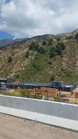 This video captures a view of a construction site nestled against a mountainous backdrop. The scene is set in Chimgan, Uzbekistan, indicated by the location context. The time of day appears to be daytime, with the sun casting shadows and illuminating the sky, which is dotted with fluffy white clouds. The weather seems to be pleasant and sunny, contributing to the overall bright and clear atmosphere.

The construction site is the central subject, featuring various equipment and structures. Prominent objects include a yellow crane, a blue truck, and several large concrete barriers that line the foreground, suggesting a road or pathway adjacent to the site. There are also what appear to be modular buildings or storage containers, fenced off with barbed wire, indicating security measures. Yellow machinery, possibly a bulldozer or similar construction vehicle, is also visible. The surrounding landscape is characterized by a steep, dry, and vegetated mountainside, with scattered trees and shrubs. The terrain suggests a somewhat arid environment.

The activity taking place is ongoing construction, evidenced by the presence of heavy machinery and building materials. The overall impression is one of development and progress in a scenic, natural setting. No people are clearly discernible in the footage, and there are no explicit emotional cues conveyed. The video does not contain any visible text.