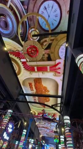 The video shows the inside of a brightly lit indoor amusement park. The ceiling is covered with an LED screen displaying a moving steampunk-themed animation with gears, pipes, and clocks. Below the screen are various rides and attractions, including a carousel and a miniature Eiffel Tower lit in blue. People are walking around and enjoying the park. The lighting is colorful, with decorative poles illuminated with vertical stripes of light. The overall impression is of a lively and whimsical environment.