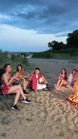 A group of seven people are enjoying themselves around a bonfire on a sandy beach in the evening. The group is composed of five women and two men. The men are standing. One man is wearing a white shirt and light blue shorts, the other a patterned shirt and jeans with rips. Both are holding drinks and engaged in conversation. Two of the women are sitting on a blanket by the fire, while three are seated in chairs slightly further away. All of them are holding drinks. The fire is blazing with flames, and there are some low-lying trees and dune grass in the background. The sky is a mix of blues and grays, suggesting either dawn or dusk. The overall atmosphere is relaxed and jovial, with music playing.