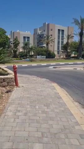 A gray SUV drives by a sidewalk with a red fire hydrant and a stone wall. In the background are apartment buildings, palm trees, and a fountain. The sky is blue and clear.