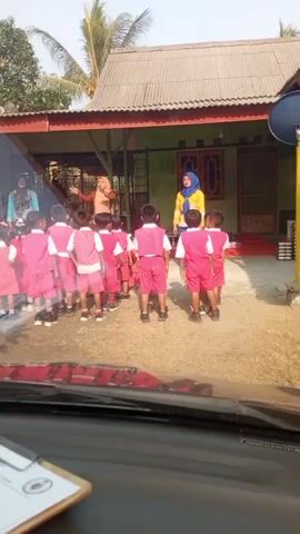 The video shows a group of young children in pink uniforms standing in a line outdoors, facing a building. Three adult women, presumably teachers, are standing in front of the children. One woman, wearing a blue hijab and yellow shirt, stands to the right, facing the children. Another woman, wearing an orange hijab and dress, stands in the center, facing the children and gesturing. A third woman, wearing a blue head covering and a patterned top, stands to the left, also facing the children. The children appear to be engaged in some sort of activity or lesson. The environment appears to be a schoolyard or outdoor area, with a building behind the group. A portion of a car interior is visible in the foreground, suggesting the video was filmed from inside a vehicle. The lighting suggests it is daytime, likely in the morning or afternoon.