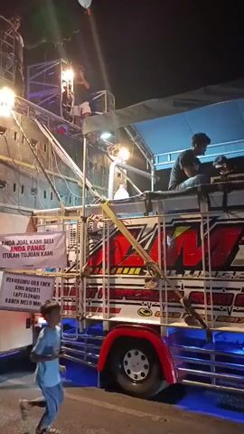 A truck is outfitted with a large sound system, with multiple speakers stacked high on a metal frame. Several people are visible on and around the truck, some appearing to operate the equipment. Two banners are displayed on the side of the truck; one reads "ANDA JUAL KAMI BELI ANDA PANAS ITU LAH TUJUAN KAMI" and the other displays a message in what appears to be Javanese script. Blue LED lights illuminate the lower part of the truck. A young person runs across the foreground of the frame. The scene is lit by artificial lights and appears to be taking place at night.