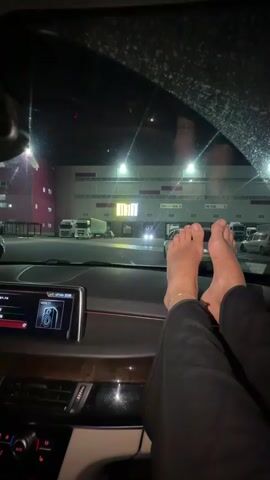 The video shows a person's bare feet resting on the dashboard of a car at night. The car is parked in front of a large building with illuminated signs and several parked trucks. The scene is dark, with artificial lights illuminating the building and surrounding area. The interior of the car is dimly lit, with some of the dashboard lights visible. The video appears to be taken from inside the car, looking out through the windshield.