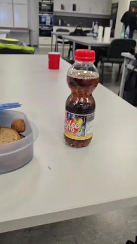 The video shows a close-up of a white table in what appears to be an office or break room. On the table is a clear plastic lunch container with a blue lid. Inside the container are several meatballs and a slice of bread. To the right of the lunch container is a bottle of iced tea with a red cap. In the background, there is a red cup, a jacket with reflective stripes, and various office equipment including cabinets and desks. The camera pans slightly to the left, revealing a tube of blue-capped substance, possibly paint or glue, also on the table. The overall scene depicts a simple lunch setup.