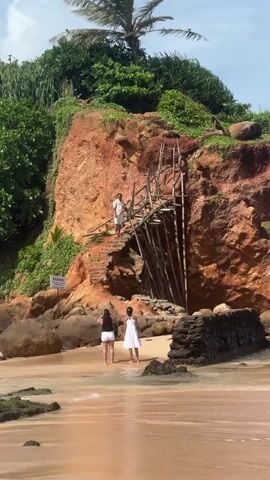 The video displays a sunny beach scene with a small, vegetated island or peninsula featuring a reddish-brown cliff. A rustic wooden and brick staircase is built into the cliff, leading down to the sandy shore. A "DANGER" sign is visible on the lower left of the cliff. One man in a light shirt and shorts is descending the staircase. On the beach directly below, two women are standing, one in a dark top and white shorts, the other in a white dress. In the foreground, two other men are standing on the sand, observing the scene; one wears a blue t-shirt and shorts, the other a patterned shirt and shorts. The sky is blue with scattered white clouds, and calm ocean waves gently lap the shore.