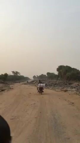 The video shows a dirt road with piles of debris and trash on either side. A person on a motorbike with a large, white bag strapped to the back is traveling down the road. There are a few trees scattered along the sides of the road, and the sky is hazy. The person on the motorbike is the main subject of the video, and their movement is the primary action.