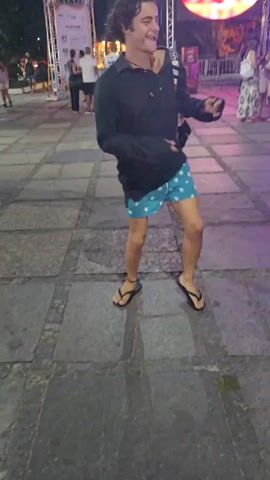 A young man dances with a smile in an outdoor square. The square is paved with gray cobblestones. The man wears a long-sleeved black shirt, light blue shorts with white stars, and black flip-flops. He has a black bag slung across his chest. A person wearing a blue tank top and patterned shorts walks past him. A clear plastic bottle lies on the ground near the bottom left corner.  In the background, there is a stage with a large circular light and a screen displaying writing in Portuguese. People stroll and stand around the square. A bicycle leans against a wall.  Streetlights and stage lights illuminate the scene.