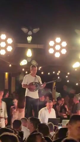 A man stands on a platform playing a stringed instrument while a crowd watches him. He is wearing a white t-shirt and dark pants. He is illuminated by stage lights and string lights. The crowd appears to be in a dimly lit outdoor venue.