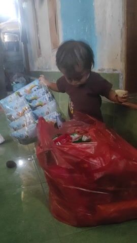 A child in a dark t-shirt and light-colored shorts is picking through a large red plastic bag filled with various items. The child appears to be holding a small, light-colored object in their right hand. The child is on their knees next to the bag, which is placed on the floor. To the left of the child, there are several items wrapped in clear plastic. The floor is a light green color. The background shows a wall with some peeling paint and a doorframe. There is also a blue basket to the left of the child. The scene appears to be indoors, in a somewhat cluttered environment.