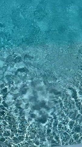 The video shows a close-up view of clear blue water, likely in a swimming pool or spa. Bubbles are rising from the bottom of the frame, indicating some form of agitation or aeration. The light refracts through the water, creating shimmering patterns on the surface and the pool floor. The perspective is from above, looking down into the water, suggesting the viewer is at the edge of the pool. No people or other objects are visible.