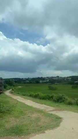 The video displays a rural landscape, likely viewed from a moving vehicle. The scene is dominated by vast green fields, characteristic of rice paddies, stretching towards a distant horizon. In the background, there are clusters of buildings, suggesting a village or small settlement, nestled among rolling hills covered in dense greenery and trees. The sky above is partly cloudy, with patches of blue visible through the white and grey clouds.

In the foreground, a metal road barrier or guardrail is visible, indicating the presence of a road from which the video is being filmed. A tall light pole stands on the left, and a signpost with unreadable text is visible on the right side of the frame in the initial seconds. As the vehicle moves, more trees and dense vegetation fill the view. The overall impression is one of a verdant, agricultural area under a moderately bright sky. No people are visible in the video. The location context is Dak Ro Wa, Vietnam, which aligns with the rural, agricultural scenery depicted.