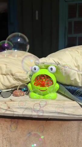 A green frog-shaped bubble machine sits on a wooden surface. It is actively blowing bubbles. The machine is between two large, light-colored cushions. The background is a dark, blurred area that appears to be outdoors. Bubbles are visible in the air, some reflecting light and colors.