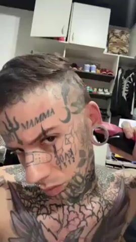 The video features a heavily tattooed man cutting his hair with a trimmer. He is shirtless and has a nose ring and stretched earlobes. His face is covered in tattoos, including the word "MAMMA" on his forehead and a drawing of a band-aid on his nose. The background is a cluttered closet with clothes, shoes, and other items. A television is visible in the background, playing an unrelated program. He is looking directly at the camera.