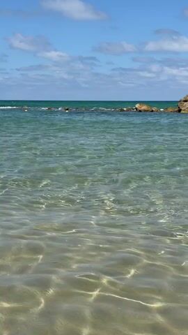 Here is a description of the video:

The video shows a clear, tranquil ocean on a sunny day. The water is shallow and sparkles in the sunlight. A line of rocks sits in the distance near the horizon. There are only a few small clouds in the blue sky.