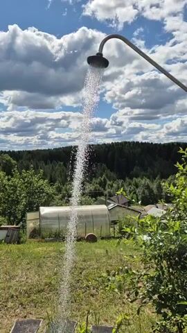 A showerhead attached to a curved metal pipe is spraying a stream of water downwards. The background consists of a grassy area with some trees, a greenhouse, and houses. The sky is blue with white, fluffy clouds. The video shows the water stream from different angles, with the showerhead and water spray remaining the central focus.