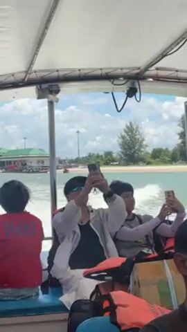 From inside a moving boat, several passengers take photos of the shoreline with their phones. The boat has a white canopy and is moving away from a building and beach with pine trees. The passengers wear life vests. The sky is partly cloudy.