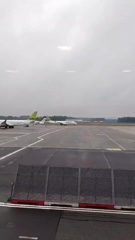 A daytime, high-angle outdoor shot shows an airport tarmac with several planes under a cloudy sky. The tarmac is dark gray and marked with white and yellow lines. A metal fence with red and white striped panels is in the foreground. Two large airplanes with light green tailfins are visible on the left, parked. A smaller white plane with light green markings is moving on the right, driving toward the camera. In the distance, a long runway stretches across the frame, with more planes parked further away. The sky is overcast and bright, with some reflections of indoor lighting visible at the top of the frame.