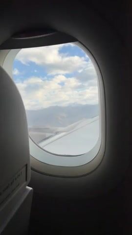 The video shows a view from an airplane window. The window frame is dark and slightly out of focus. Through the window, a vast mountainous landscape is visible. The mountains are covered in snow on their peaks, and the lower slopes appear rocky and barren, with some evidence of dry vegetation or earth. Clouds are scattered across a bright blue sky. The wing of the airplane is visible in the lower right portion of the frame, extending into the landscape. The camera appears to be handheld and slightly shaky, with the view panning slowly across the scene. The light suggests it is daytime, possibly late afternoon or early morning due to the soft lighting on the mountains.